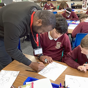 Yr artist Mfikela Jean Samuel yn helpu disgybl yn y dosbarth
